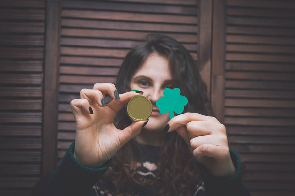young beautiful woman with painted green clover on her cheek holding golden coin at the bar in front of a wooden wall - Foto, afbeelding
