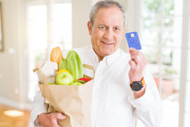 Handsome senior man holding paper bag full of fresh groceries and showing credit card as payment - Foto, Imagem