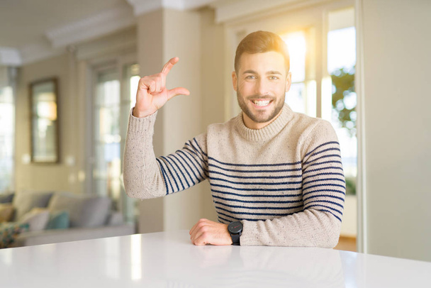 Young handsome man at home smiling and confident gesturing with hand doing size sign with fingers while looking and the camera. Measure concept. - Photo, Image