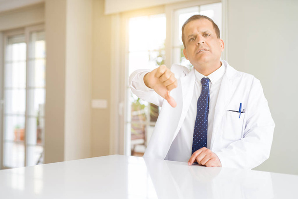 Středního věku lékař muž na sobě lékařský plášť na klinice, nešťastný a rozzlobený, zobrazeno odmítnutí a negativní s palce dolů gesto. Chybný výraz. - Fotografie, Obrázek