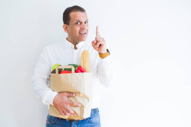 中年男持株食料品ショッピング アイデアに驚いて白い背景の上にバッグや幸せそうな顔、ナンバーワンと人差し指を質問 - 写真・画像