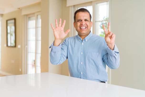 Homem de meia idade sentado em casa mostrando e apontando para cima com os dedos número sete, sorrindo confiante e feliz
. - Foto, Imagem
