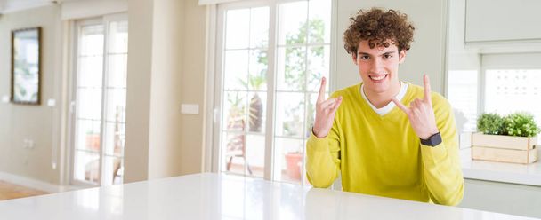 Wide angle shot of young handsome man at home shouting with crazy expression doing rock symbol with hands up. Music star. Heavy concept. - Fotografie, Obrázek