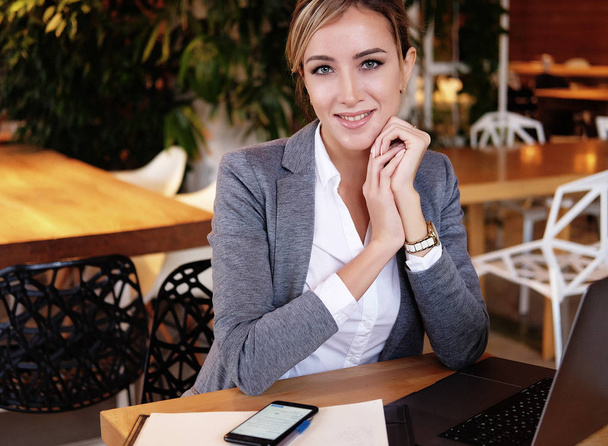 Young businesswoman sitting at desk and working. Smiling and looking  at camera. Lifestyle, people and business concept. - Zdjęcie, obraz