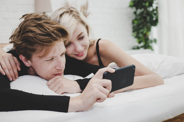 Przystojny chłopak trzymając smartfon i oglądając film ze swoją dziewczyną w łóżku. Cute para poczuć relaks. Miłośnicy atrakcyjny, ciesząc się ich życie i dostać szczęście uśmiech w wakacje w domu. - Zdjęcie, obraz