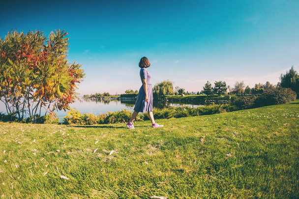 Chica delgada caminando a lo largo del río. Una mujer en un vestido está caminando sobre la hierba verde. Morena está descansando en el parque
. - Foto, imagen