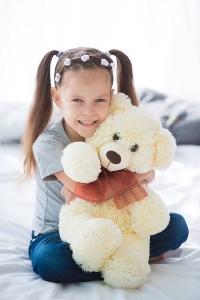 Adorable smiling littlegirl sitting on a bed hugging a white teddy bear. - Photo, Image