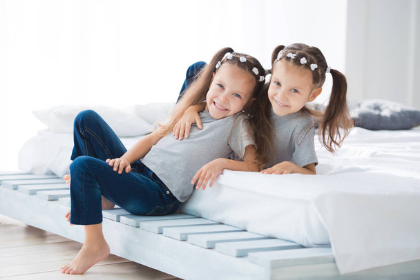 Two little cute smiling girls sisters twins are sitting on the bed in the room. - Foto, Imagem