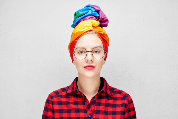 A pretty young woman with round glasses and full lips in a rainbow turban and a red shirt is smiling against a white wall. LGBTQ Lesbian, gay, bisexual, transgender, queer. Homosexual man - Zdjęcie, obraz
