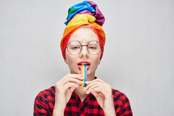 A pretty young woman with round glasses and full lips in a turban and a red shirt holds a rainbow in her mouth against a white wall. LGBTQ Lesbian, gay, bisexual, transgender, queer. Homosexual man - Foto, immagini