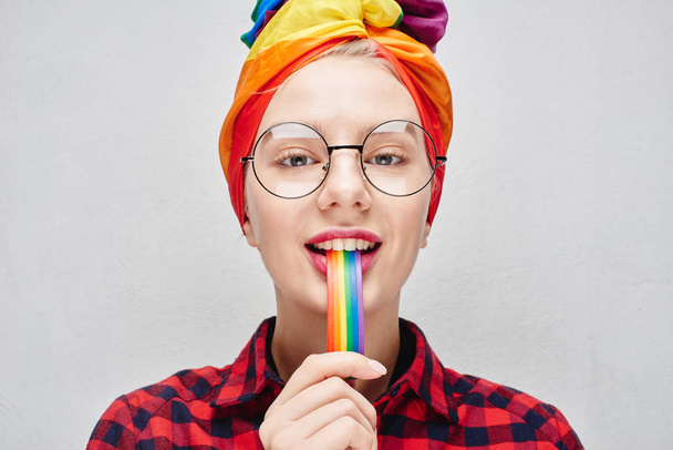 A pretty young woman with round glasses and full lips in a turban and a red shirt holds a rainbow in her mouth against a white wall. LGBTQ Lesbian, gay, bisexual, transgender, queer. Homosexual man - Φωτογραφία, εικόνα