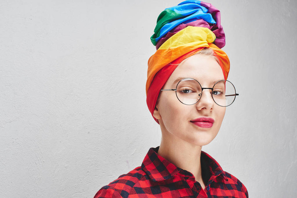 A pretty young woman with round glasses and full lips in a rainbow turban and a red shirt is smiling against a white wall. LGBTQ Lesbian, gay, bisexual, transgender, queer. Homosexual man - Foto, Imagen