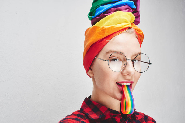 A pretty young woman with round glasses and full lips in a turban and a red shirt holds a rainbow in her mouth against a white wall. LGBTQ Lesbian, gay, bisexual, transgender, queer. Homosexual man - 写真・画像