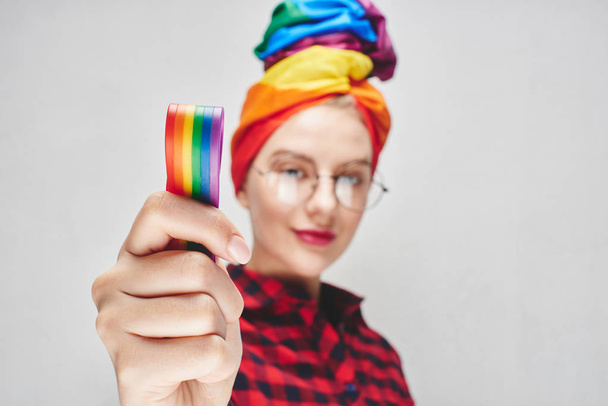 A beautiful young woman with round glasses and full lips in a turban and a red shirt holds a rainbow in her hand against a white wall. LGBTQ Lesbians, gays, bisexuals, transgender, queer. Homosexual man - Zdjęcie, obraz