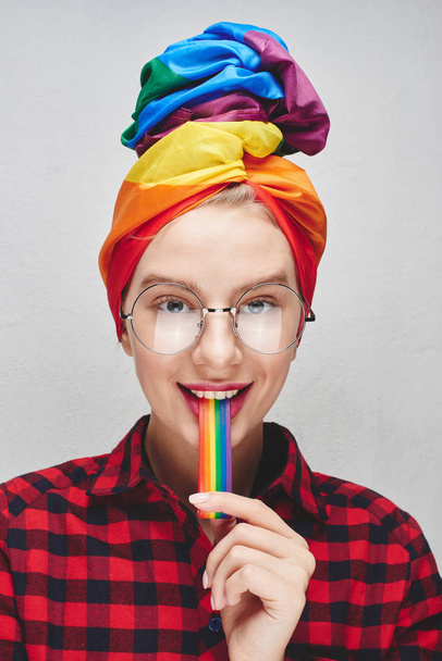 A pretty young woman with round glasses and full lips in a turban and a red shirt holds a rainbow in her mouth against a white wall. LGBTQ Lesbian, gay, bisexual, transgender, queer. Homosexual man - Foto, Bild
