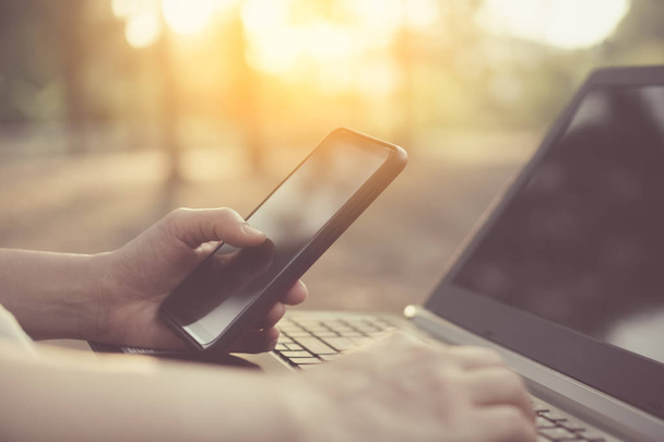 Mulher mão usando telefone inteligente e laptop no parque natural ao ar livre e céu pôr do sol com fundo abstrato luz bokeh
. - Foto, Imagem