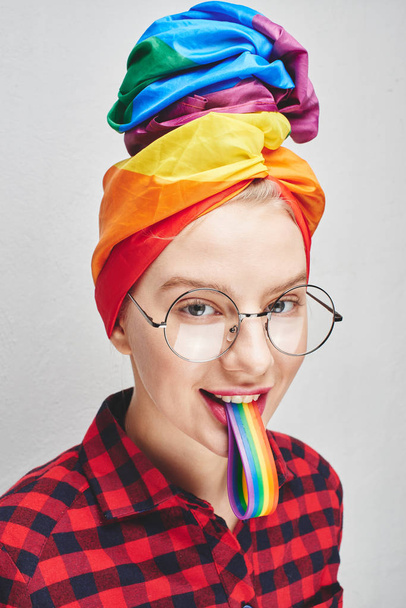 A pretty young woman with round glasses and full lips in a turban and a red shirt holds a rainbow in her mouth against a white wall. LGBTQ Lesbian, gay, bisexual, transgender, queer. Homosexual man - Фото, зображення