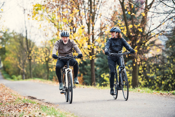 A senior couple with electrobikes cycling outdoors on a road in park in autumn. - Fotografie, Obrázek