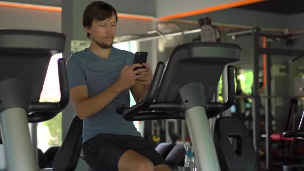 A young man at the gym on an exercise bike holding the phone in front of his eyes in his hands. All the attention is in the phone. The concept of dependence on social networks. Mobile addiction - Кадри, відео