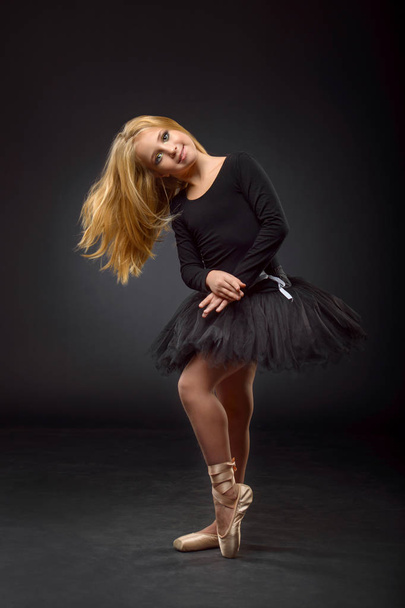 Cute little ballerina with long hair in a black tutu and pointe posing on a black background - Foto, Imagen