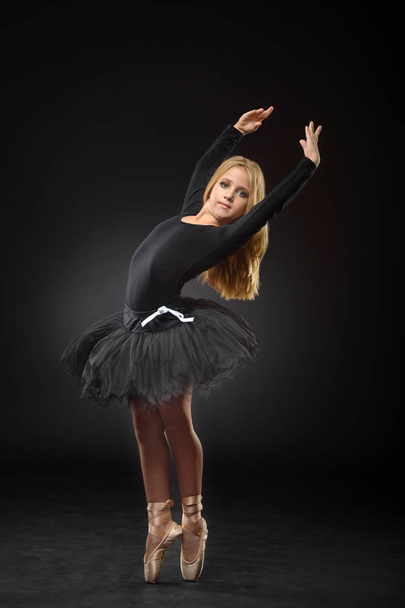 Cute little ballerina with long hair in a black tutu and pointe posing on a black background - Φωτογραφία, εικόνα