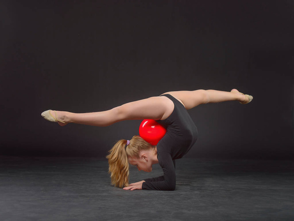 Young beautiful white caucasian girl gymnast doing gymnastic exercise with a red sports ball on black background - Foto, Imagem
