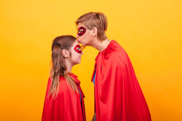 Mutter im Superman-Kostüm küsst sein kleines Mädchen auf die Stirn vor gelbem Hintergrund - Foto, Bild