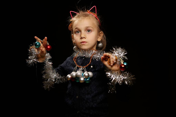 Κορίτσι παιδί ηλικίας 5-7 ετών ντυμένος ως ένα χριστουγεννιάτικο δέντρο, εξετάζει το πλαίσιο, το συναισθηματικό πορτραίτο στούντιο το νέο έτος σε μαύρο φόντο - Φωτογραφία, εικόνα
