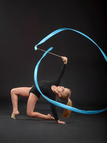 Young beautiful white caucasian girl gymnast doing gymnastic exercise with blue ribbon on black background. - Foto, imagen