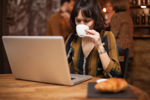 Брюнетка деловая женщина наслаждается чашкой чашки во время чтения электронной почты на ноутбуке
 - Фото, изображение