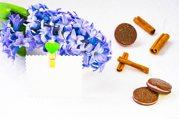 Flower composition. Blue hyacinth lies in the background, a number of dishes and chocolate chip cookies, still life adorn cinnamon sticks. Nearby there is a paper card for a signature decorated with a baby doll with a heart. The background is bright. - Photo, Image