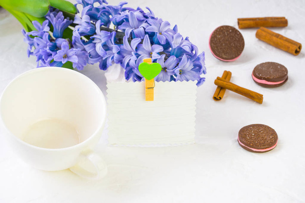 White empty mug on a background of spring flowers. Blue hyacinth lies in the background, a number of dishes and chocolate chip cookies, still life adorn cinnamon sticks. Nearby there is a paper card for a signature decorated with a baby doll with a h - Photo, Image