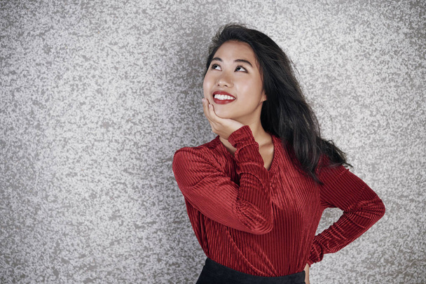 Азиатская юная брюнетка в элегантной красной блузке и красной помаде на губах улыбается на сером фоне
 - Фото, изображение