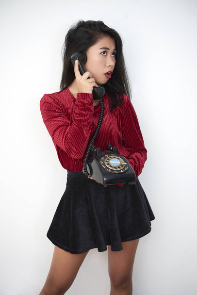 Asiática jovem mulher de pé em saia curta e falando em um telefone antiquado isolado no fundo branco
 - Foto, Imagem