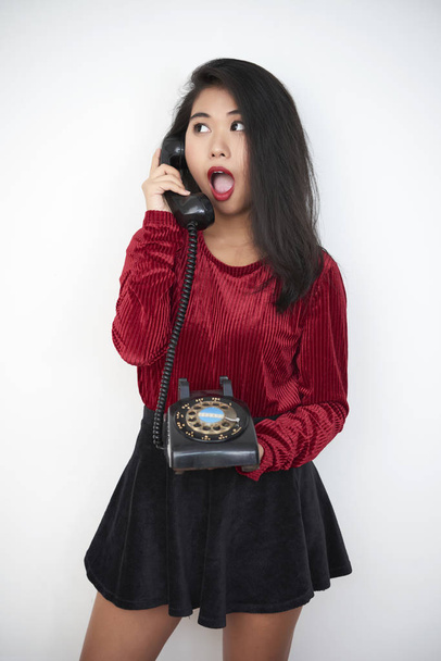 Young pretty girl standing with short skirt and talking on vintage telephone with surprised look on white background - Foto, Bild