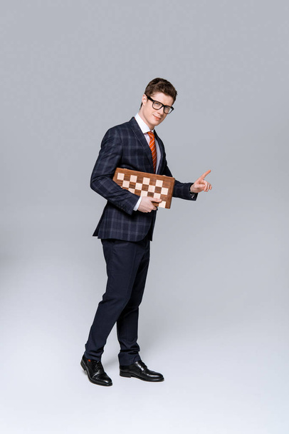 young businessman pointing and holding chess board isolated on grey  - Fotó, kép