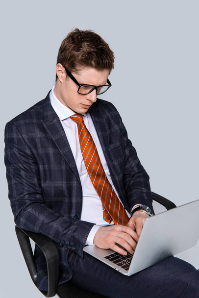young businessman sitting on chair and using laptop isolated on grey - Photo, Image
