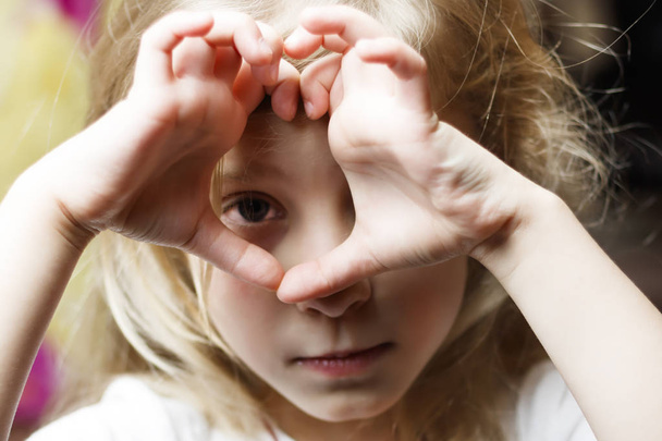 a girl with white hair makes a heart and looks into it. Concept view in childhood. there is toning - Фото, зображення