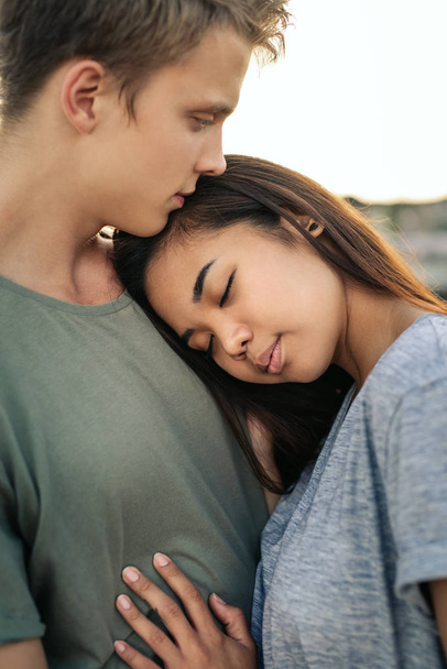 Young woman with her eyes closed hugging her boyfriend while standing arm in arm together by a harbor in the late afternoon - Fotoğraf, Görsel