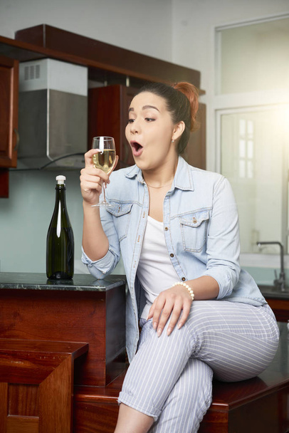 Chica joven sentada en la cocina y mirando el vaso con vino en la mano con sorpresa
 - Foto, Imagen