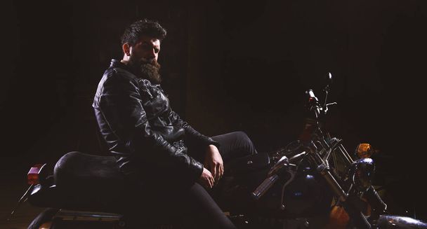 Man with beard, biker in leather jacket sitting on motor bike in darkness, black background. Macho, brutal biker in leather jacket riding motorcycle at night time, copy space. Night racer concept - Fotografie, Obrázek