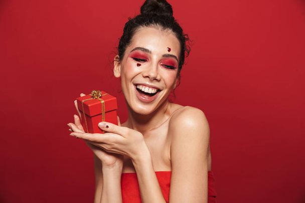 Beautiful happy cheerful young woman with red bright makeup isolated over red wall background posing with hearts on face holding gift box. - Fotó, kép