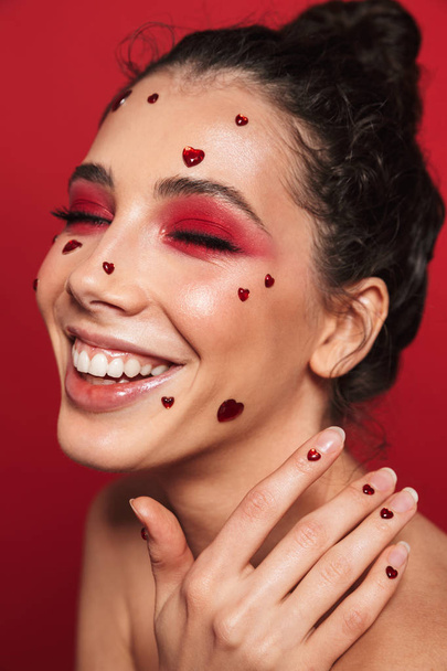 Rire jeune femme avec un maquillage lumineux rouge isolé sur fond de mur rouge posant avec des cœurs sur le visage
. - Photo, image