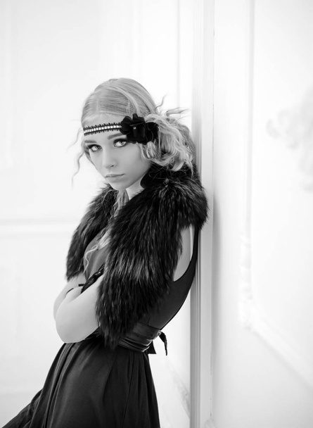 Young movie star of roaring twenties posing isolated on white background. Blond girl in headband with flower and fur collar leaning on wall - Φωτογραφία, εικόνα