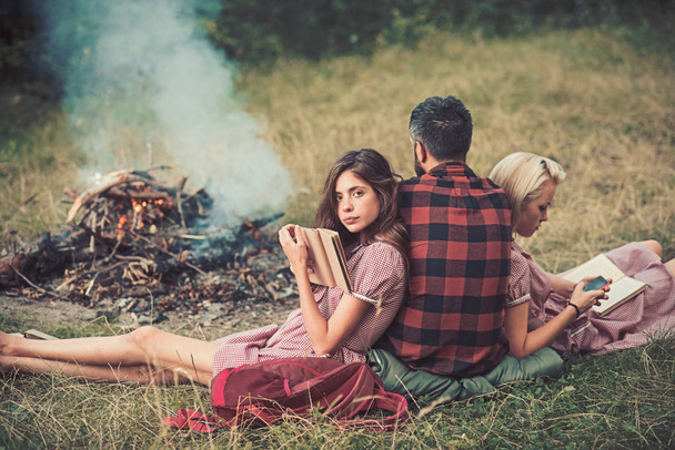 Camping in wilderness. Turn back guy looking at fire while two beautiful girls read book, sustainable education concept - Photo, image