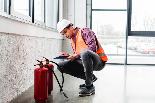 handsome fireman in helmet sitting near extinguishers and holding clipboard - Photo, Image