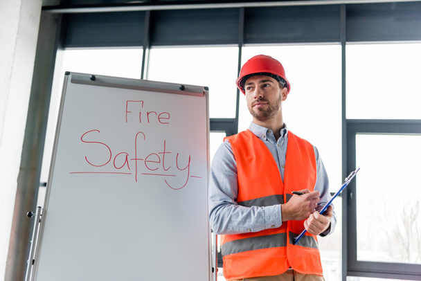 bonito bombeiro no capacete segurando prancheta e caneta enquanto estava perto de placa branca com letras de segurança contra incêndio
 - Foto, Imagem