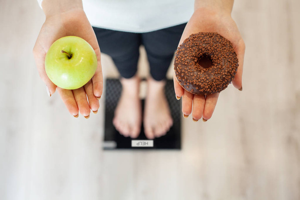 Kevyt. Nainen mittaamalla kehon paino punnitusasteikolla tilalla donitsi ja omena. Makeiset ovat epäterveellistä roskaruokaa. Ruokavalio, Terveellinen syöminen, Elämäntapa. Painonpudotus. Liikalihavuus. Ylhäältä
 - Valokuva, kuva
