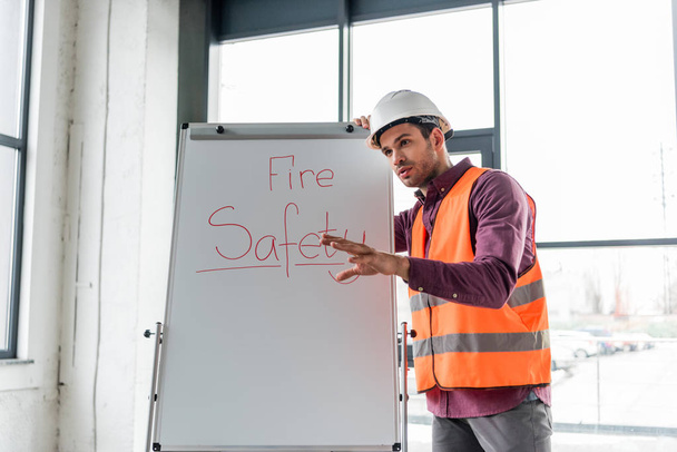 handsome firefighter in helmet talking while standing near white board with fire safety lettering - Foto, immagini
