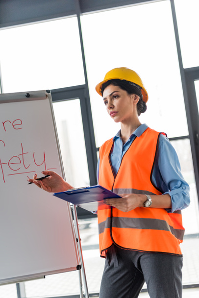 serious female firefighter in helmet holding clipboard and pen while standing near white board with fire safety lettering - Photo, image